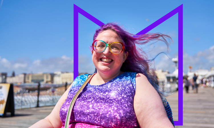 A woman in a wheelchair on a pier on a sunny day. She's smiling at the camera and her purple-dyed hair is blowing in the wind.
