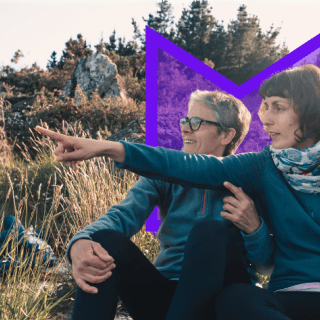 Two women sit in nature, looking to the left of frame where one of them is pointing.